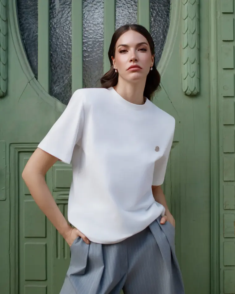 Model in weissem Oberteil und grauer weiter Hose vor einer grünen Haustür, aus der Kampagne Leap of Faith.