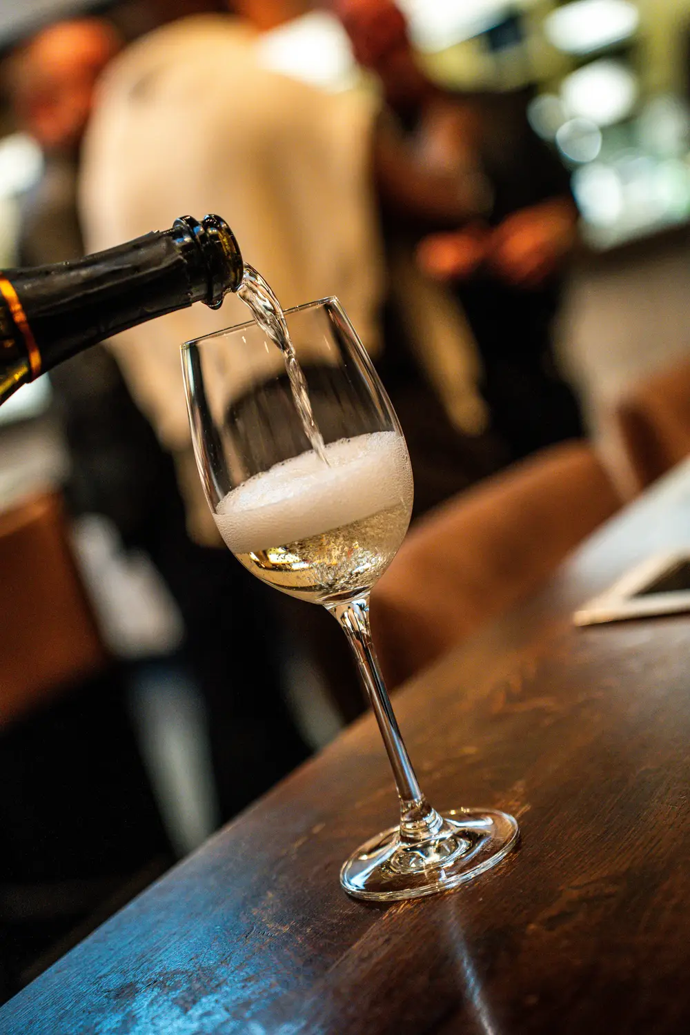 Weisswein wird in ein Glas auf einem dunklen Holztresen eingeschenkt, der Hintergrund ist unscharf.