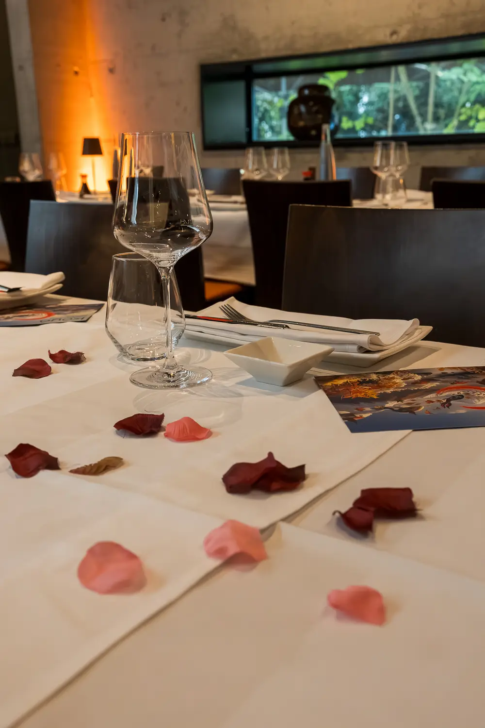Ein Gedeck aus der Nähe: gefaltete Serviette, Weinglas und Besteck auf weissem Tuch, dazwischen einzelne Rosenblätter.