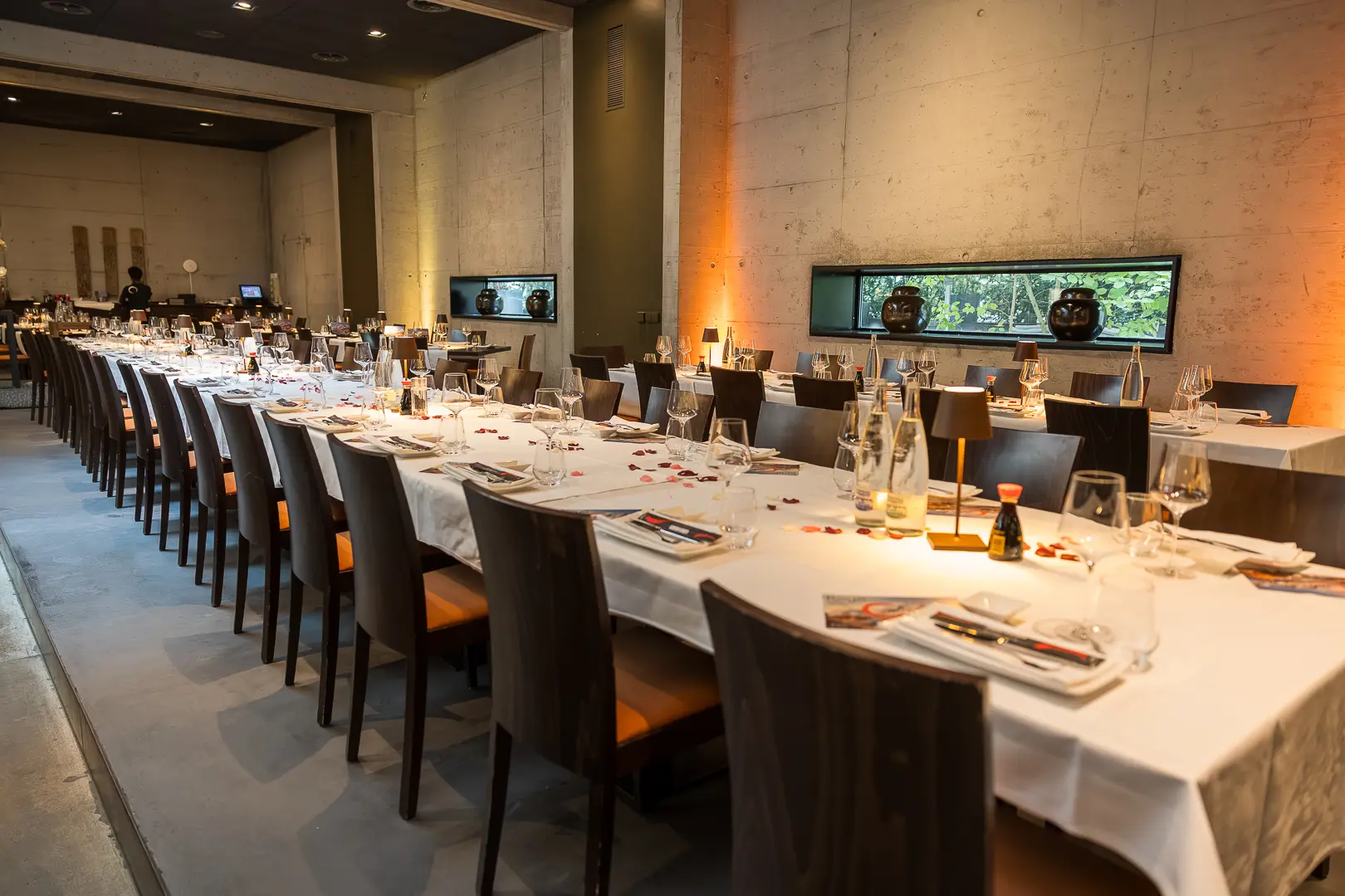 Der Saal vor dem Dinner: eine lange gedeckte Tafel zieht sich durch den Raum, dahinter eine Betonwand in warmem Licht.