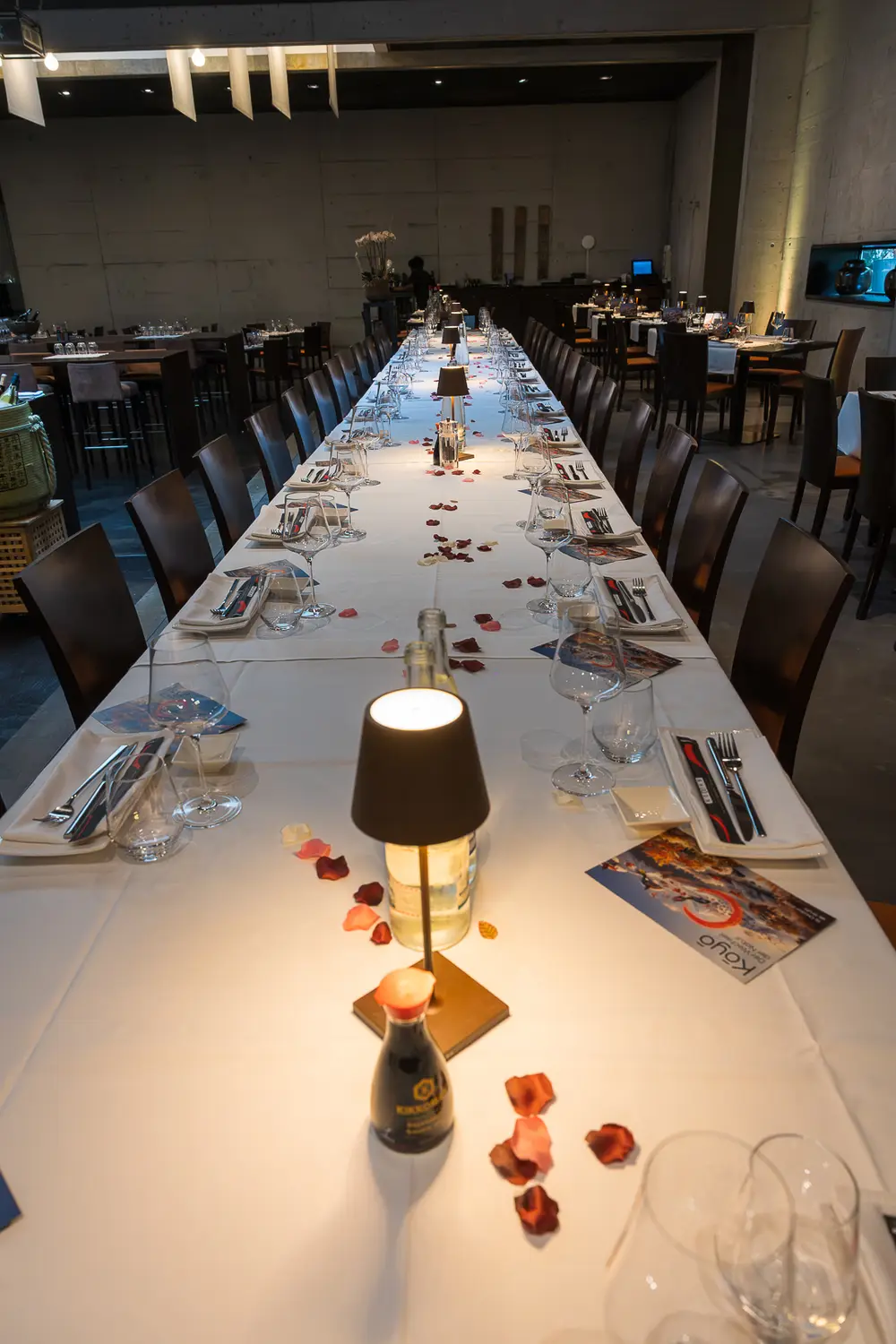 Eine Tischlampe steht auf der weissen Tafel, ringsum Rosenblätter, Besteck und Gläser, der Tisch verliert sich in der Tiefe.