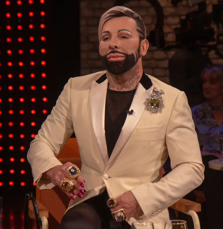 Harald Glööckler in cremefarbenem Smoking mit Brosche und Ringen, sitzend in einem Fernsehstudio.