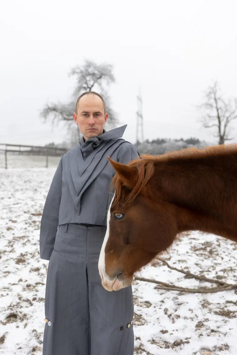 Model in einem grauen, weit geschnittenen Anzug mit drapiertem Stehkragen steht in einem verschneiten Feld, daneben der Kopf eines Fuchspferdes.
