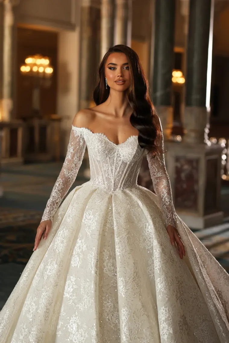 Model in einem schulterfreien Brautkleid mit Spitzenärmeln und weitem Rock in einem Saal mit Säulen.