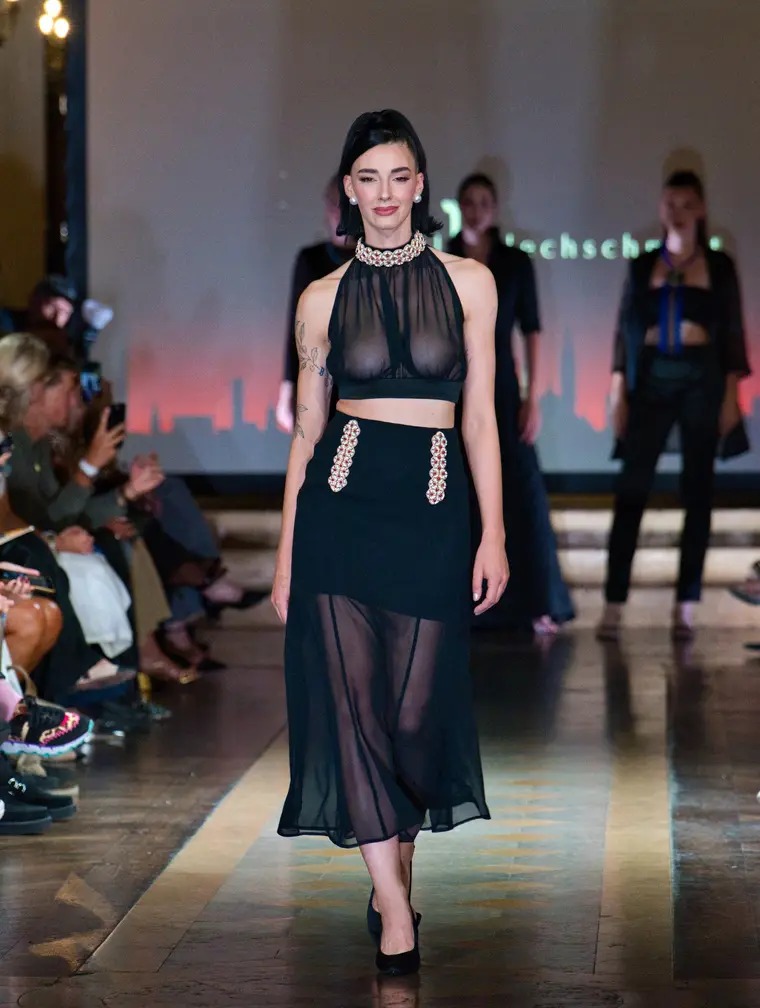 Model in einem transparenten schwarzen Oberteil und langem schwarzem Rock in einem dunklen Saal.